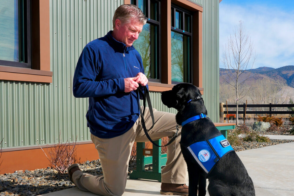 Man kneeling with a black service dog outside a building.